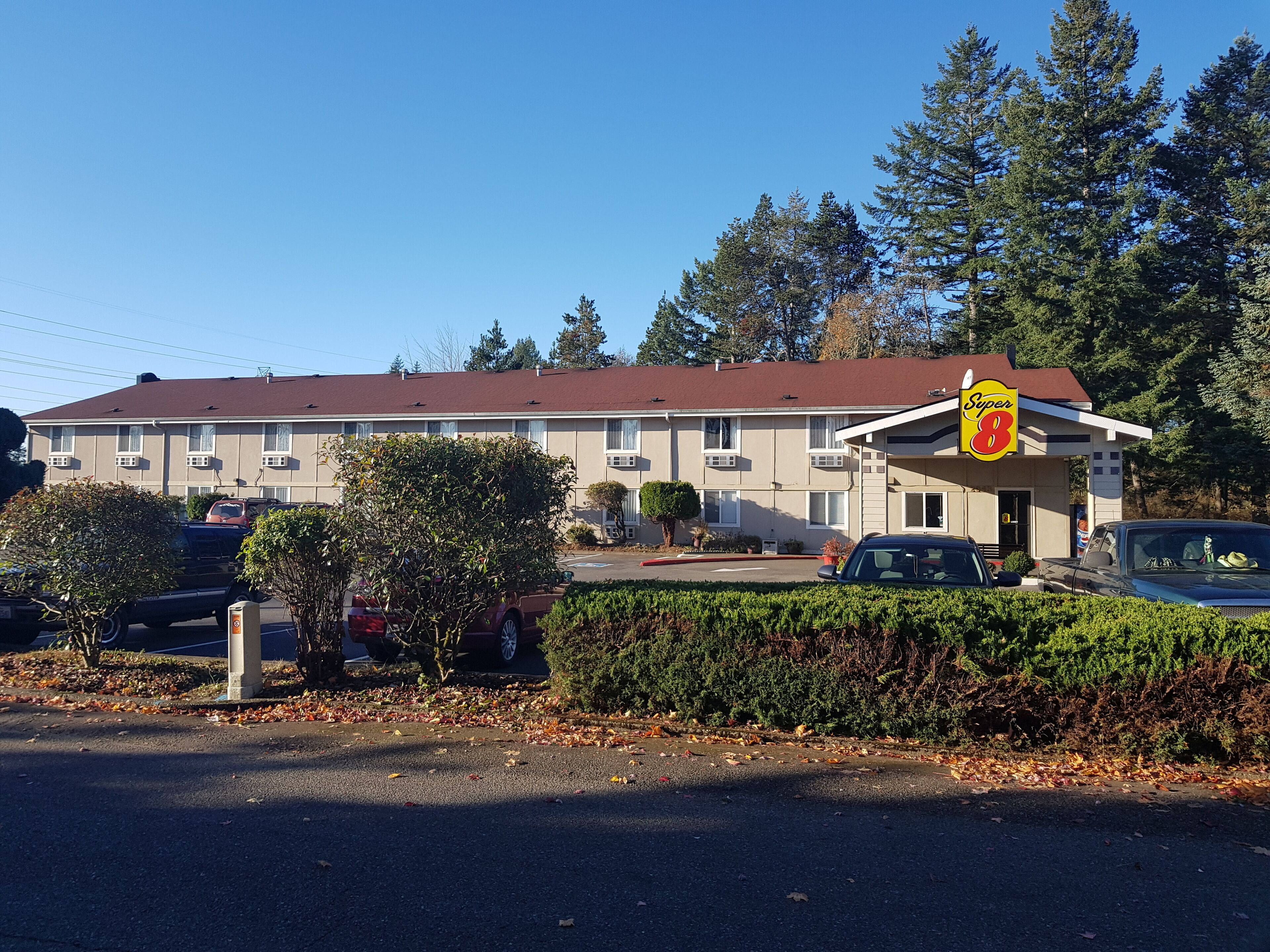 A motel with a red roof, beige exterior, and a 'Super 8' sign. There are cars parked in front, and a hedge in the foreground.