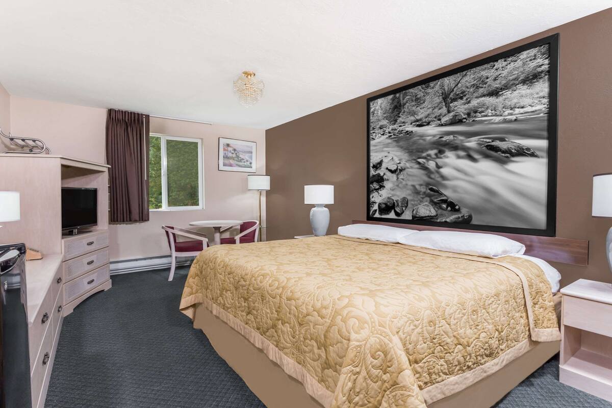Ein Hotelzimmer mit einem Bett, einem Schreibtisch mit Fernseher, einem Sessel, einem großen Fenster und einem Gemälde einer Flusslandschaft.