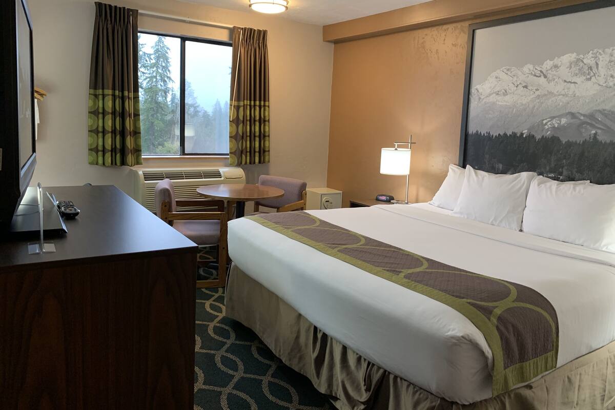 Ein Hotelzimmer mit einem Bett, einem Schreibtisch mit Computer, einem Sessel, einem Fenster mit Vorhängen, einer Wandlampe und einem Gemälde von einer Berglandschaft.