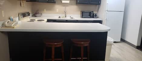 A kitchen with a white countertop, black cabinets, a microwave, a refrigerator, and two wooden stools.