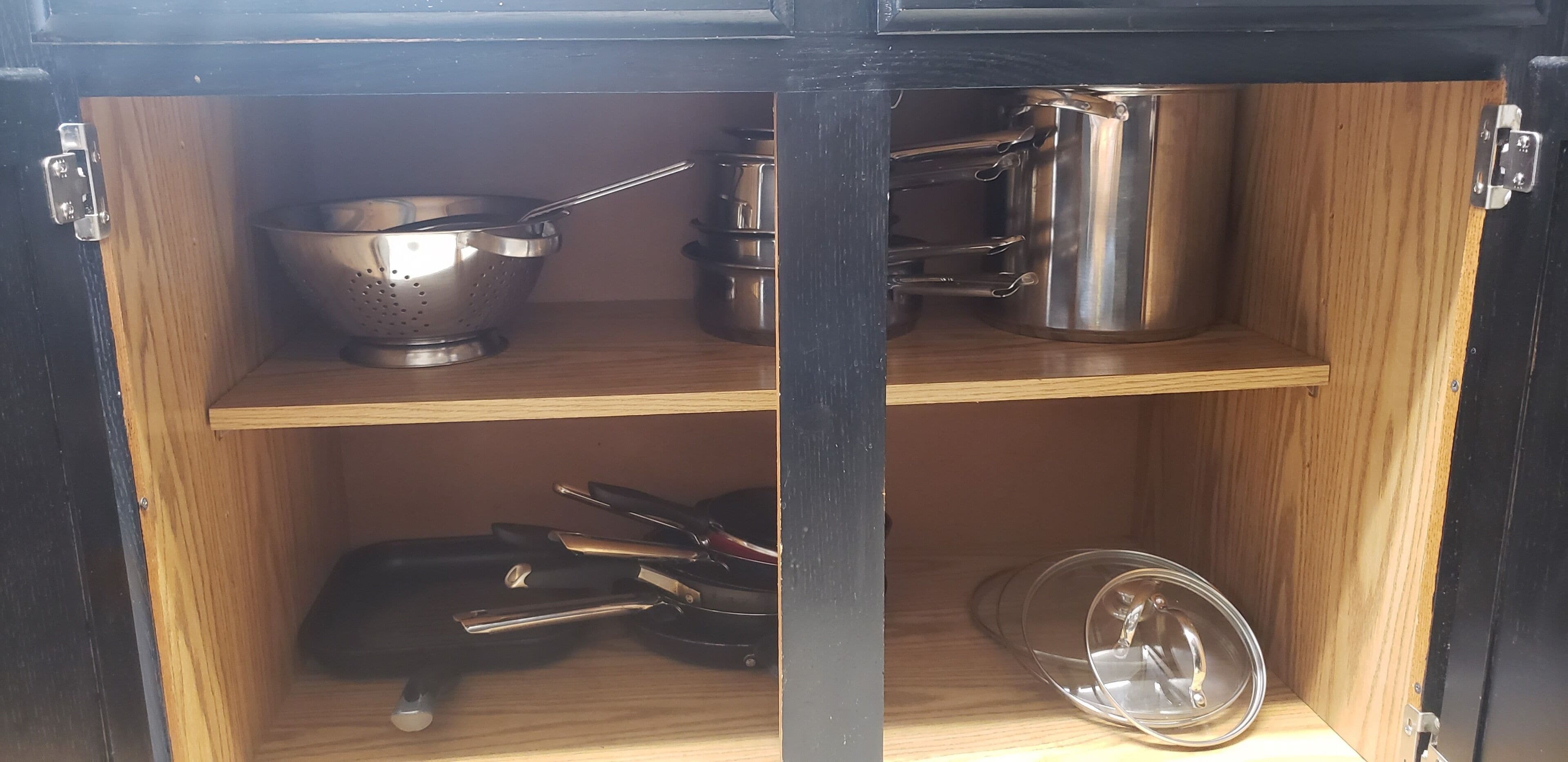 Een keukenkastje met roestvrijstalen kookgerei, waaronder pannen, braadpannen en een vergiet.