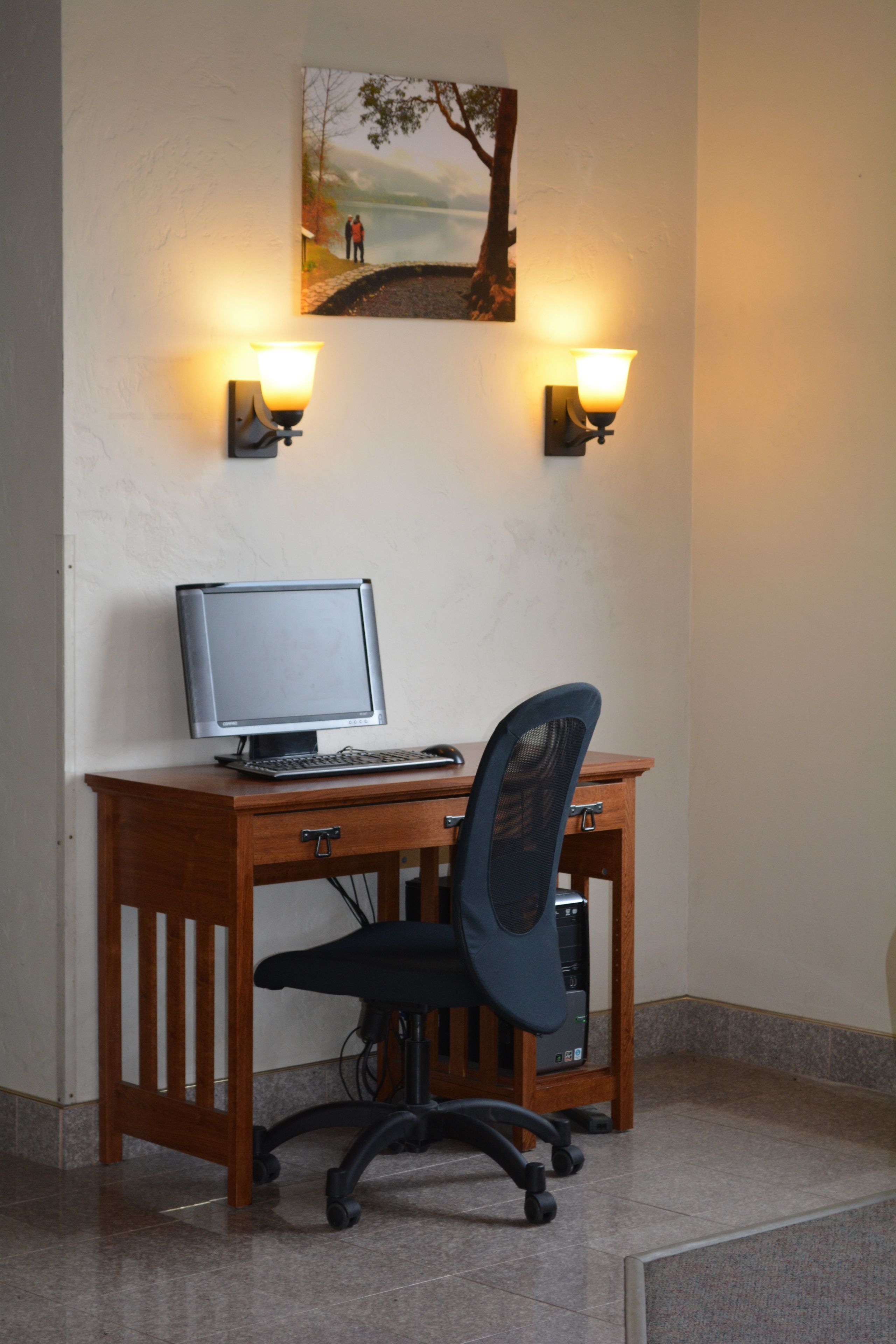 Een houten bureau met een computer en stoel, twee wandlampen en een schilderij van een boom en mensen aan de muur.