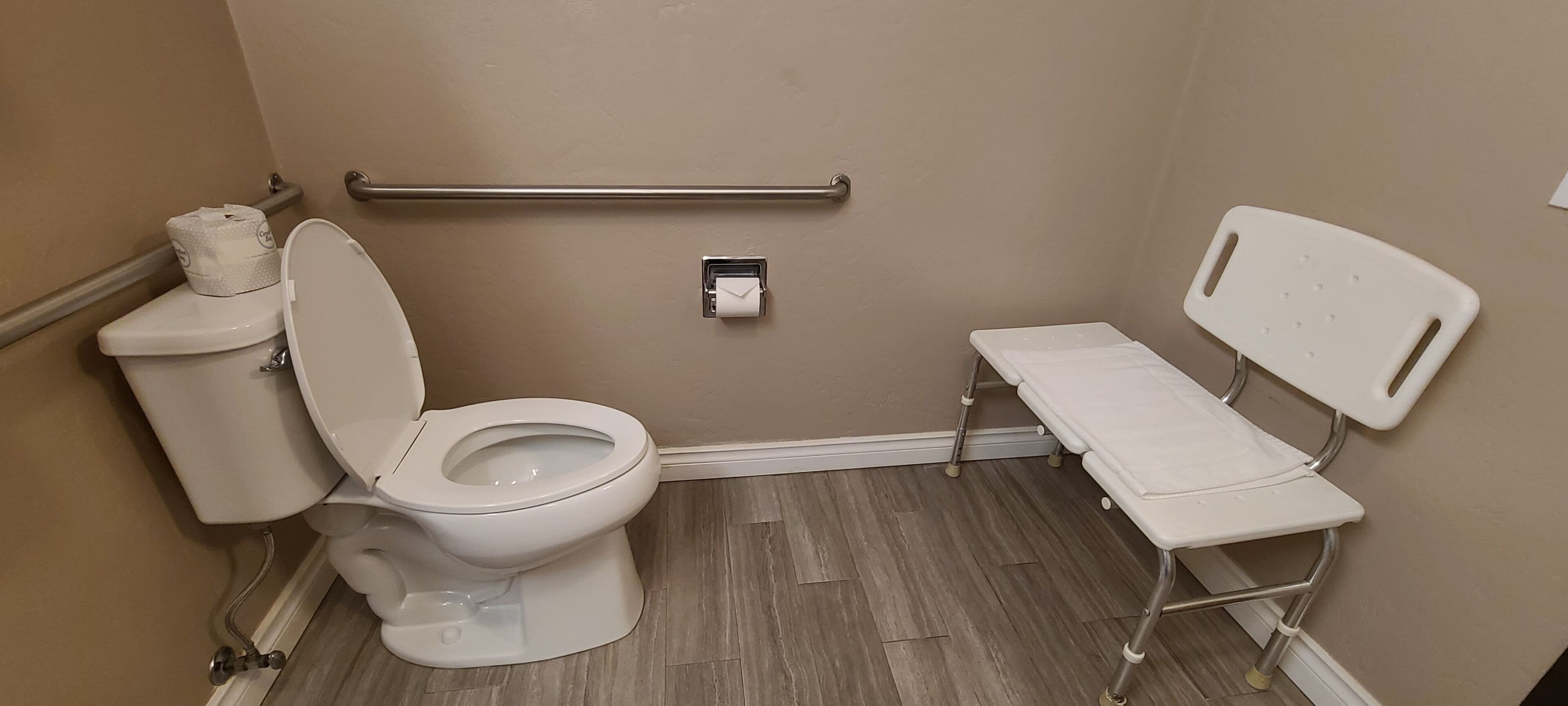 A bathroom with a toilet, a handrail, a bench, and a toilet paper holder.