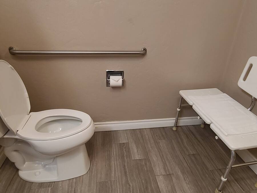 A bathroom with a toilet, a handrail, a bench, and a toilet paper holder.