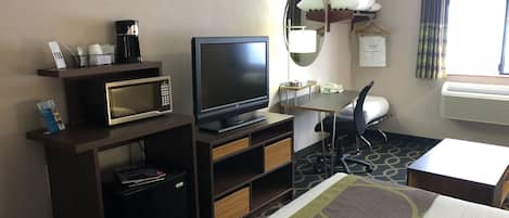 Habitación de hotel con cama, escritorio con silla, televisor, microondas, cafetera, espejo y ventana con cortinas.