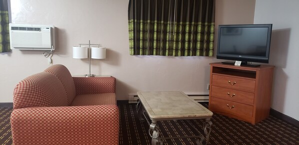 Habitación de hotel con un sofá a cuadros, una mesita, un televisor sobre un soporte de madera, una lámpara y un aire acondicionado.