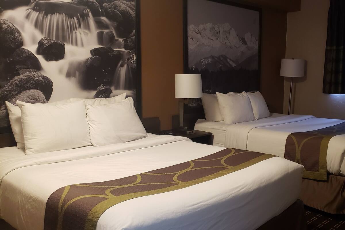 Ein Hotelzimmer mit zwei Betten, einem großen Gemälde einer Wasserfalllandschaft, einem Bergpanorama, einer Lampe und einem Fenster mit Vorhängen.
