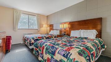 Ein Hotelzimmer mit zwei Betten, einem Holz-Kopfteil, einem Fenster mit Vorhängen, einem Schreibtisch mit Stuhl und einer Wandlampe.
