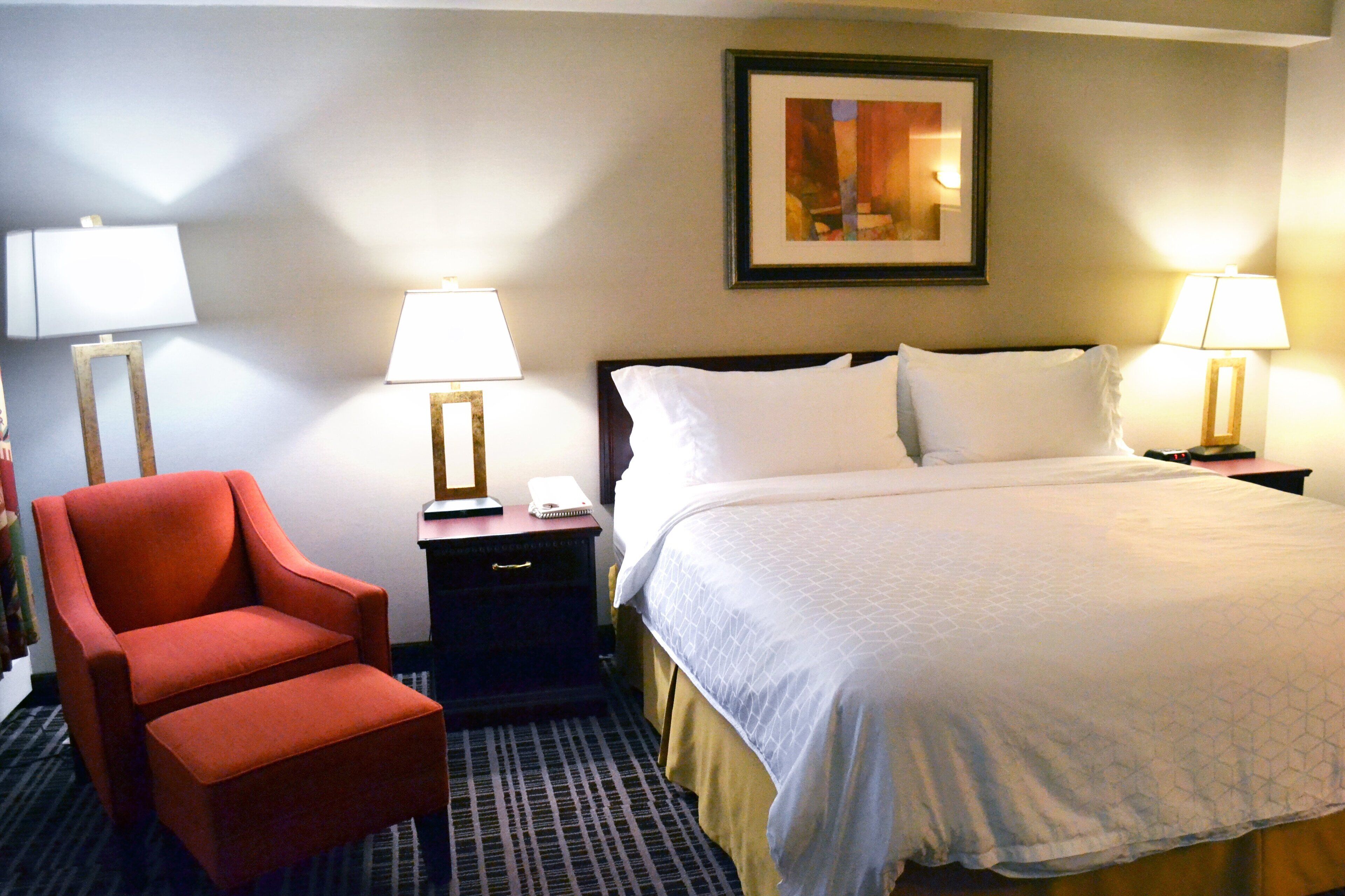 A hotel room with a bed, two bedside tables with lamps, a red armchair, and a framed picture on the wall.