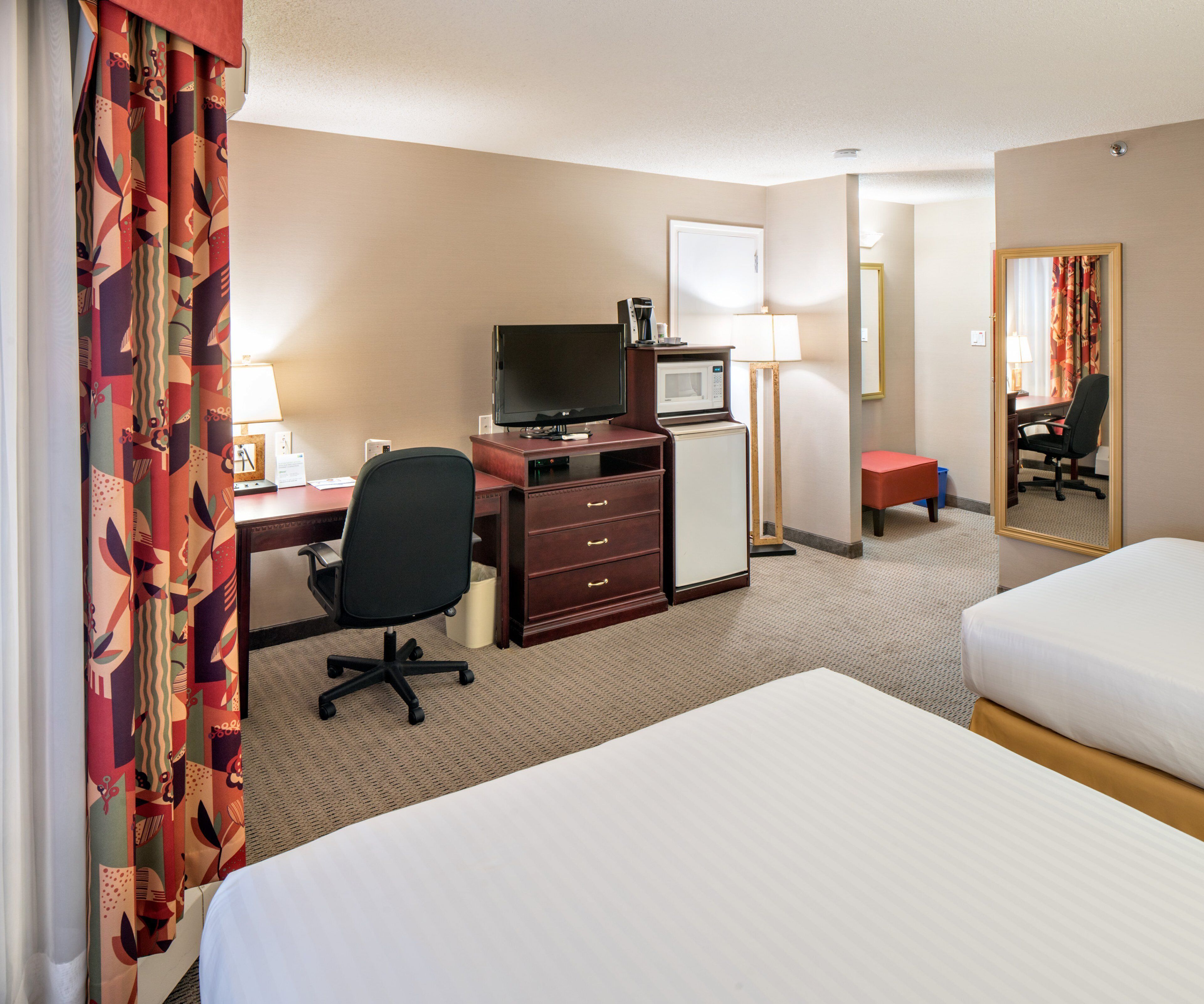 A hotel room with a bed, a desk with a computer, a TV, a microwave, and a small table with a lamp.