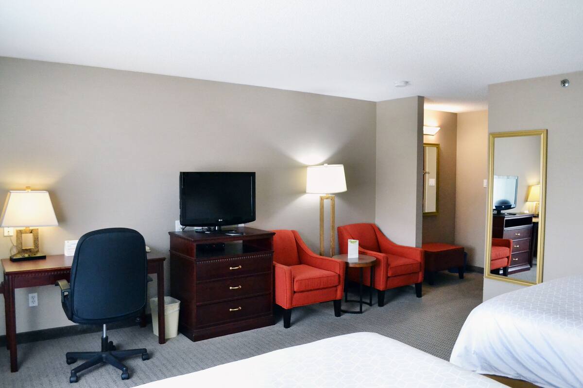 A hotel room with a bed, a desk with a computer, a television, two red armchairs, a small table, and a mirror.