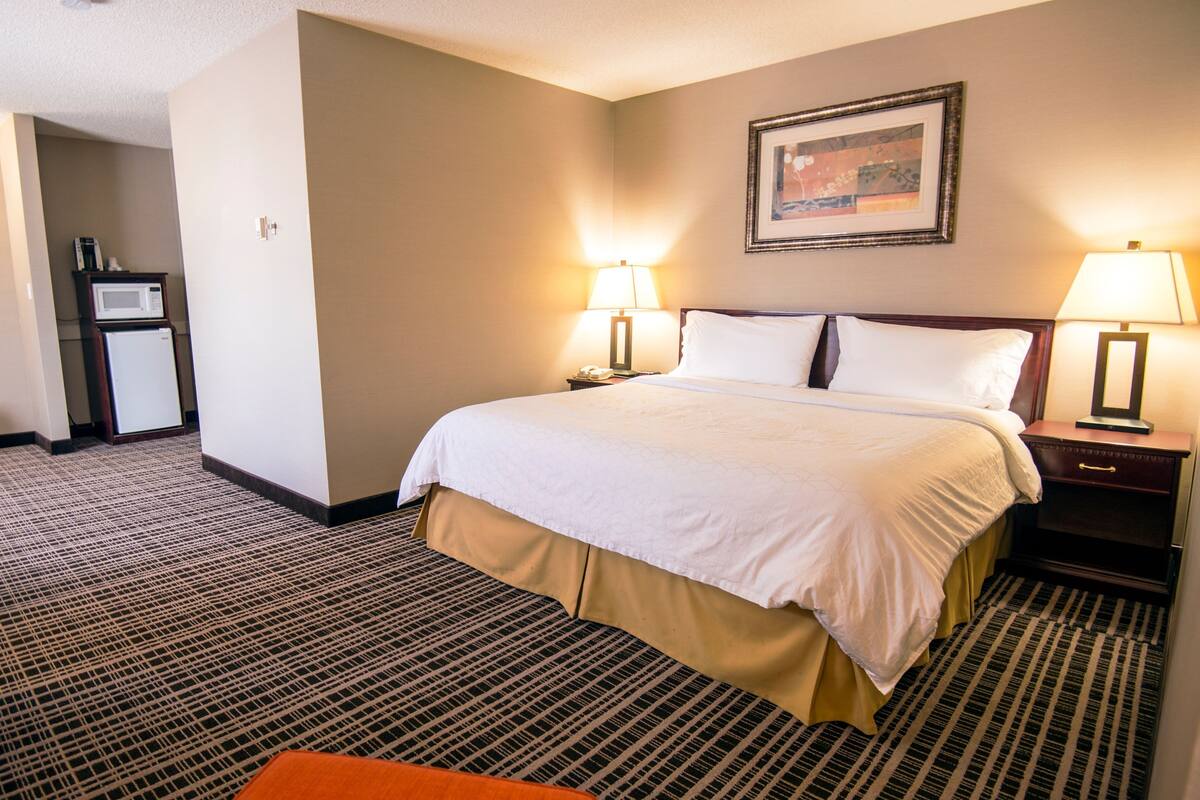 A hotel room with a large bed, two bedside lamps, a small table, a microwave, and a refrigerator.