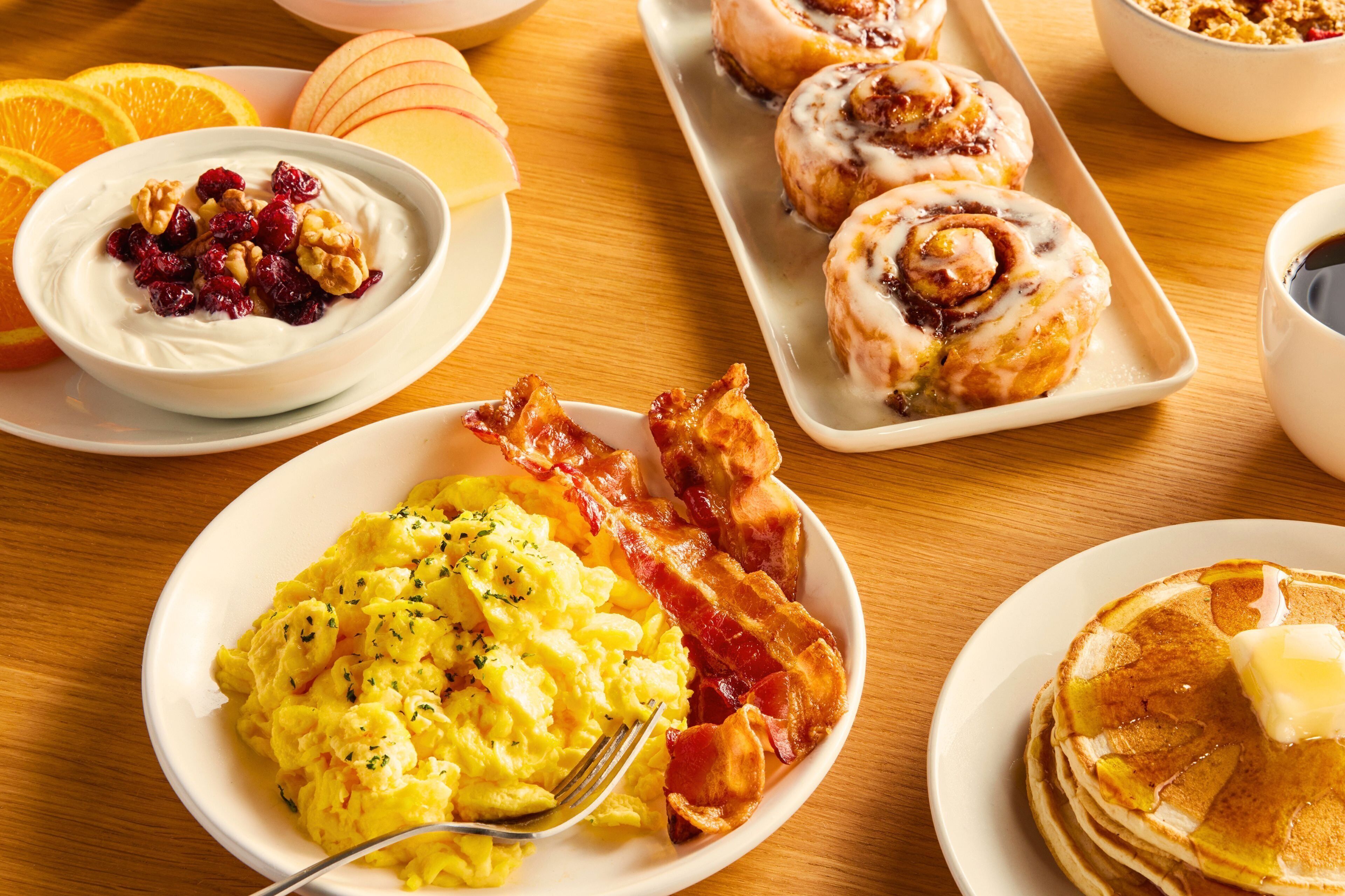 A breakfast table with scrambled eggs, bacon, pancakes, and pastries.