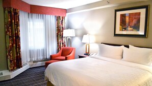 A hotel room with a large bed, a red armchair, a standing lamp, and a painting on the wall.