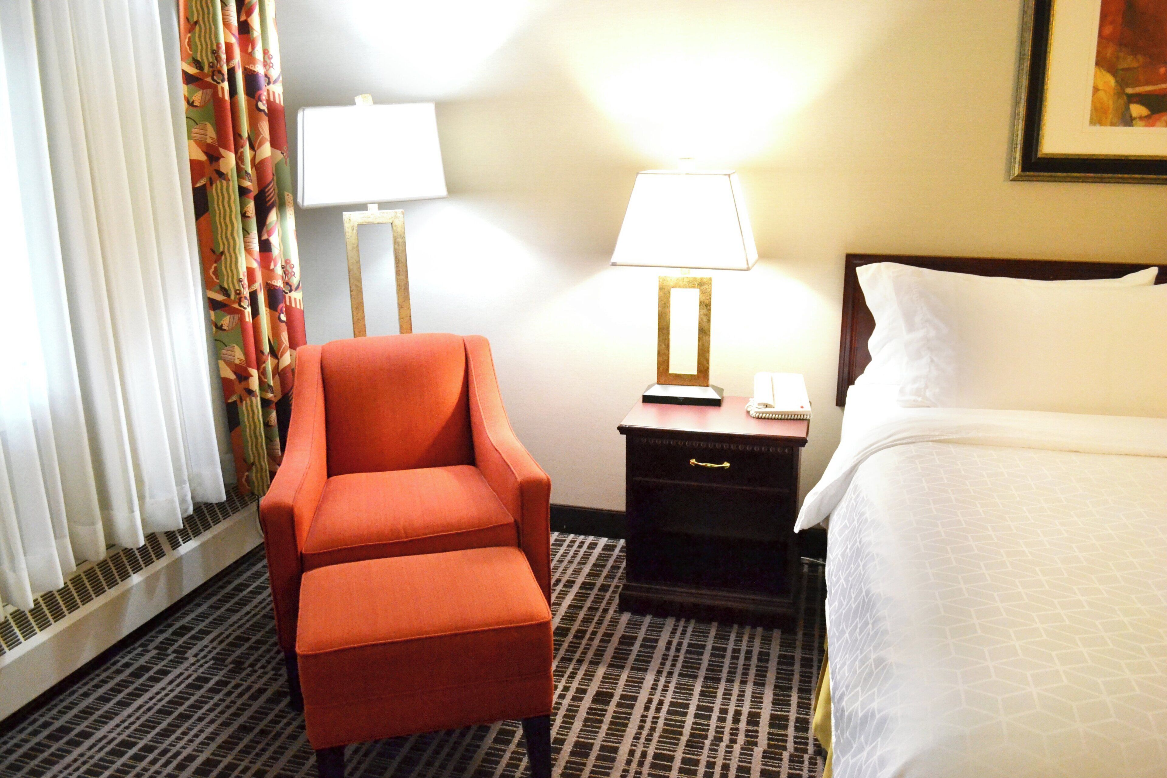 A hotel room with a single bed, a nightstand with a lamp, a chair, and a window with curtains.