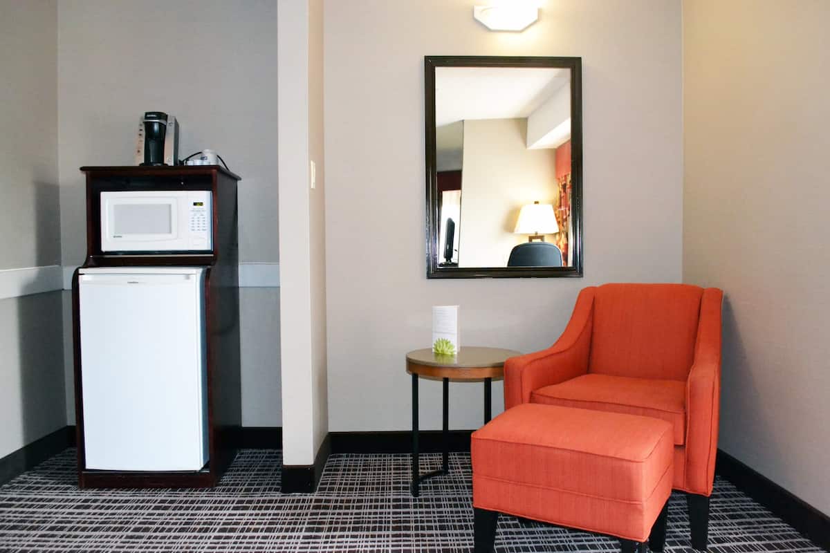 A hotel room with a compact kitchenette, a small table with a lamp, and a single orange chair.