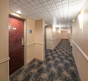 Un pasillo de hotel con alfombra estampada, una puerta de madera y un cartel azul con el número de la habitación.