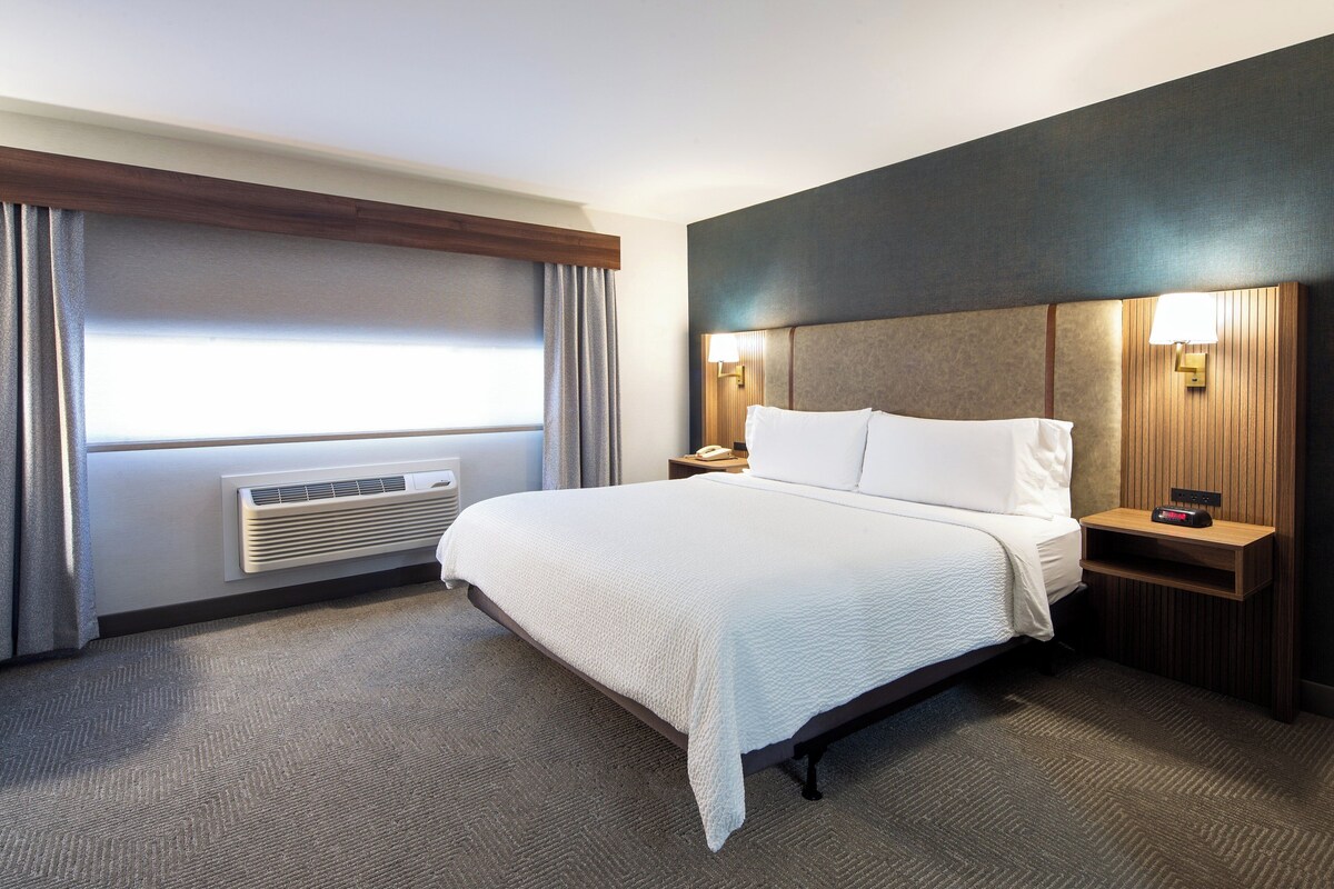 A hotel room with a large bed, a bedside table with a lamp, a wall-mounted heater, and a window with curtains.