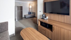 A compact hotel room with a kitchenette, a flat-screen TV mounted on a wooden panel, a seating area with a sofa and a small table, and a refrigerator. - Holiday Inn Hinton by IHG (Hinton)