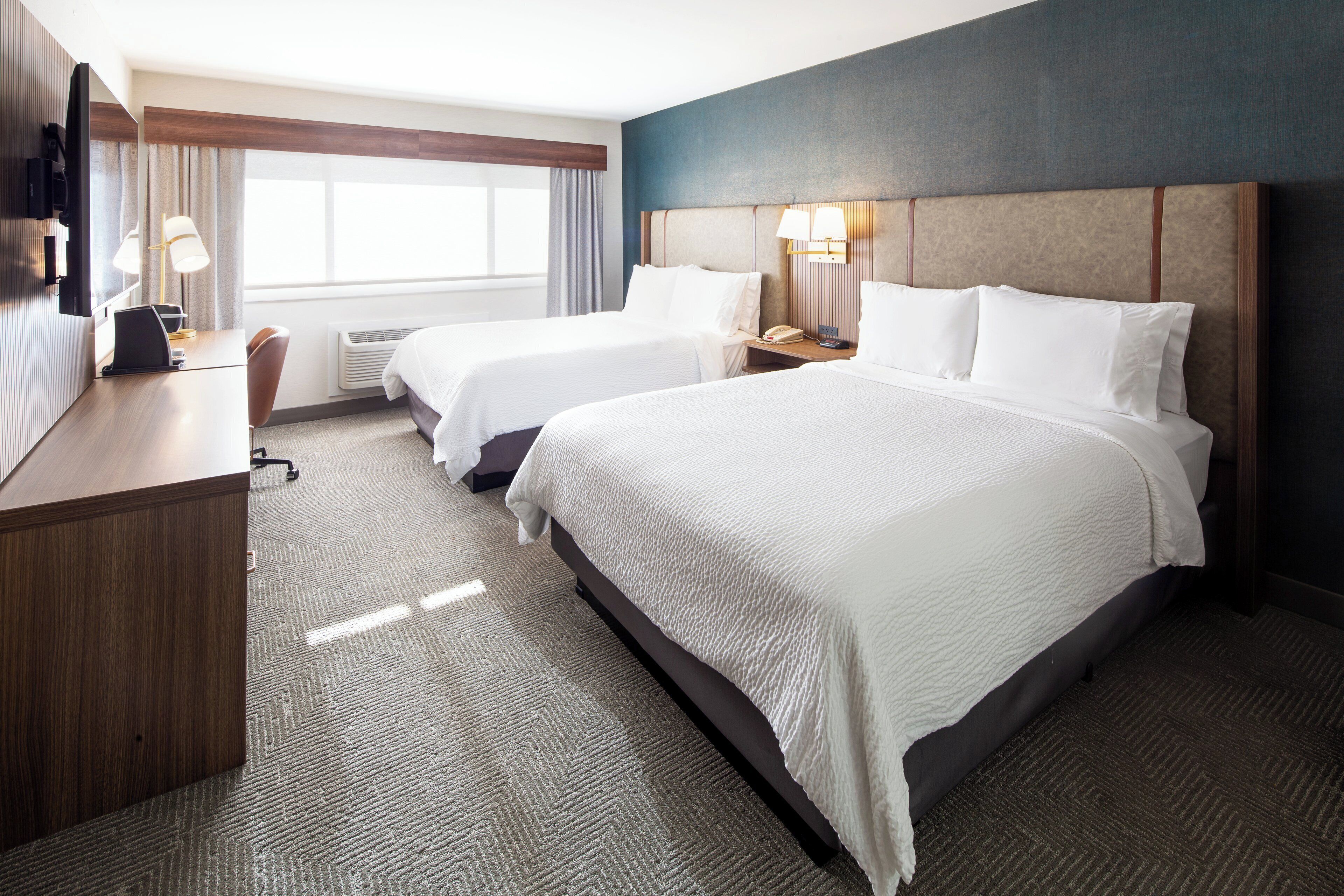 A hotel room with two beds, a desk, a chair, a lamp, and a window with curtains.