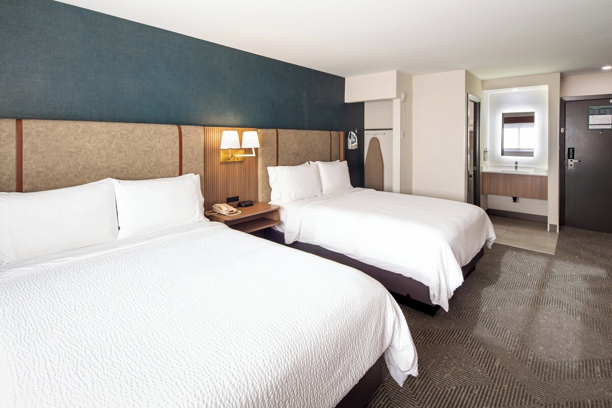 A hotel room with two beds, a nightstand with a lamp, a mirror, and a door leading to another room.