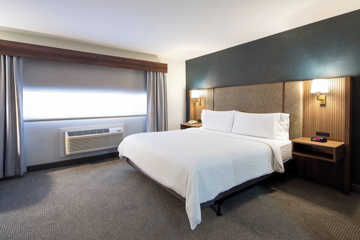 Una habitación de hotel con una cama grande, una mesita de noche con lámpara, un calentador en la pared y una ventana con cortinas.