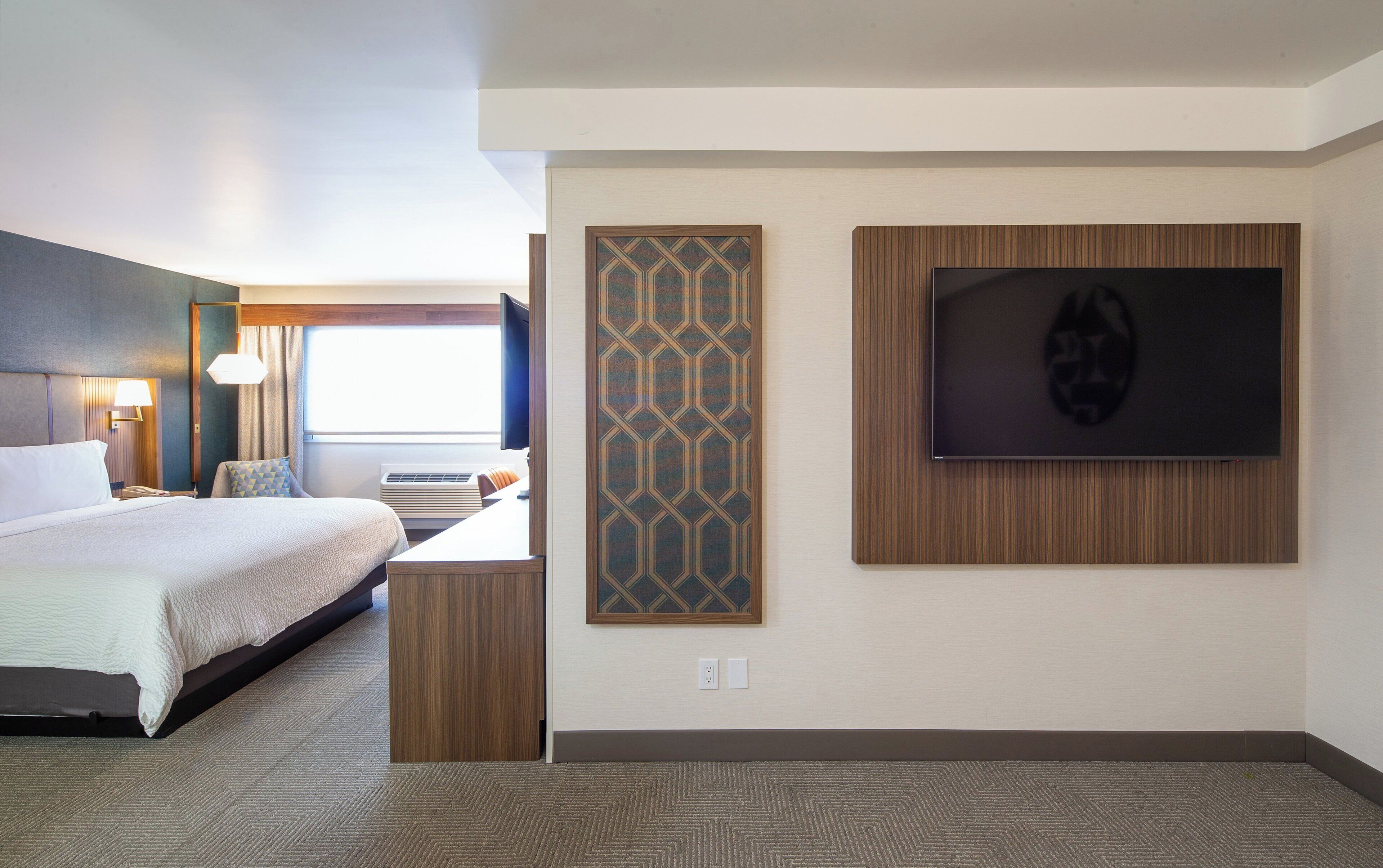 Habitación de hotel con cama, escritorio con computadora, televisor montado en la pared y decoración mural con estampado.