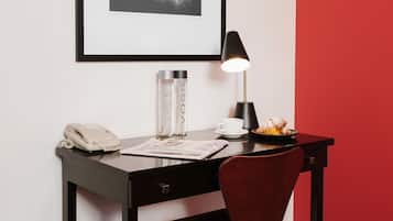 Un bureau avec un téléphone, une lampe, une tasse et une assiette contenant de la nourriture, le tout placé devant un mur rouge orné d’un tableau encadré.
