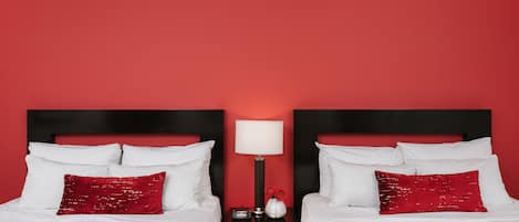 Habitación de hotel con dos camas, cada una con ropa de cama blanca y almohadas rojas. Entre las camas hay una mesita de noche con una lámpara y un jarrón con flores.