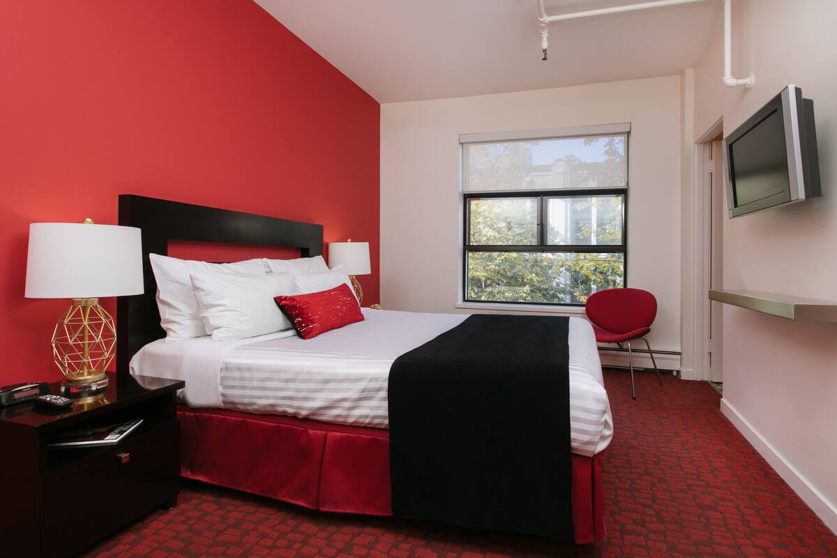 A hotel room with a red carpet, a bed with white bedding and a black headboard, a red chair, a TV mounted on the wall, and a window with a view of trees.
