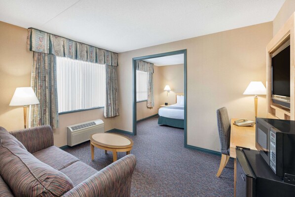 A hotel room with a sofa, a small round table, a chair, a microwave, a TV, a bed, and a bathroom visible through an open door. - Ramada Plaza by Wyndham Prince George (Prince George)