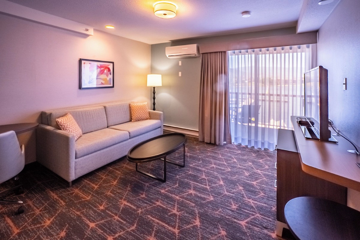 Une chambre d’hôtel avec un canapé, une petite table, un bureau et une grande fenêtre avec des stores.