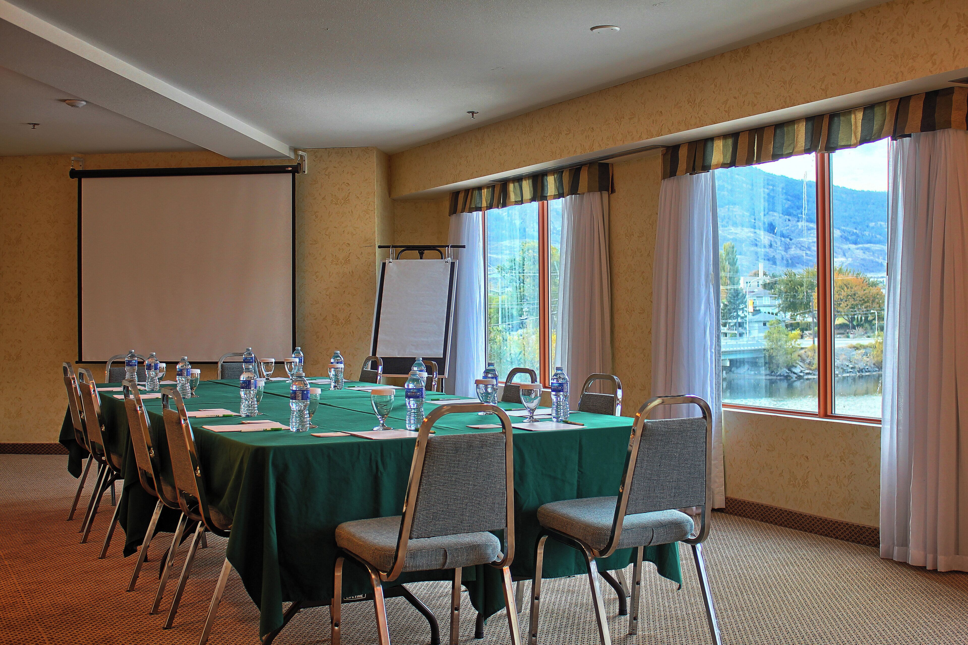 A conference room with a large table, chairs, a projector screen, and windows with curtains.