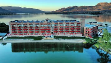 A multi-story hotel with balconies, situated by a lake with mountains in the background.