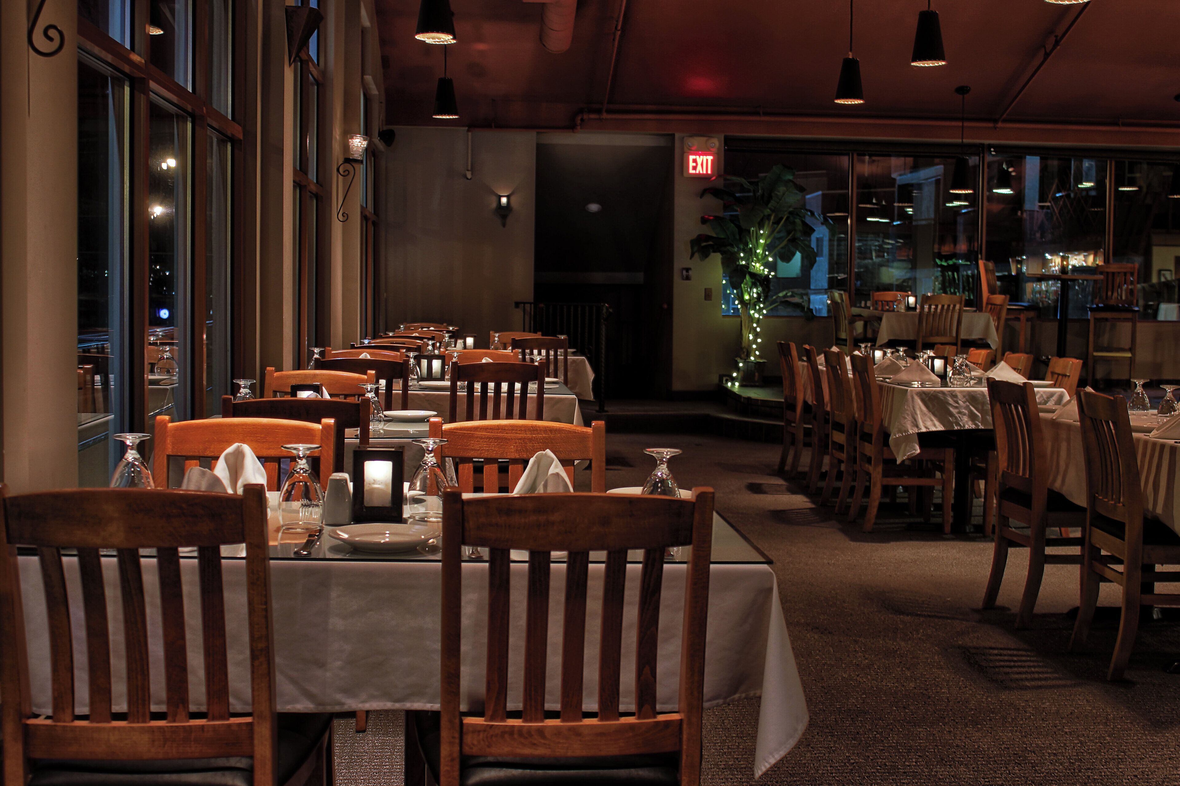 Interior de un restaurante con varias mesas y sillas, un letrero de salida visible y una planta al fondo.