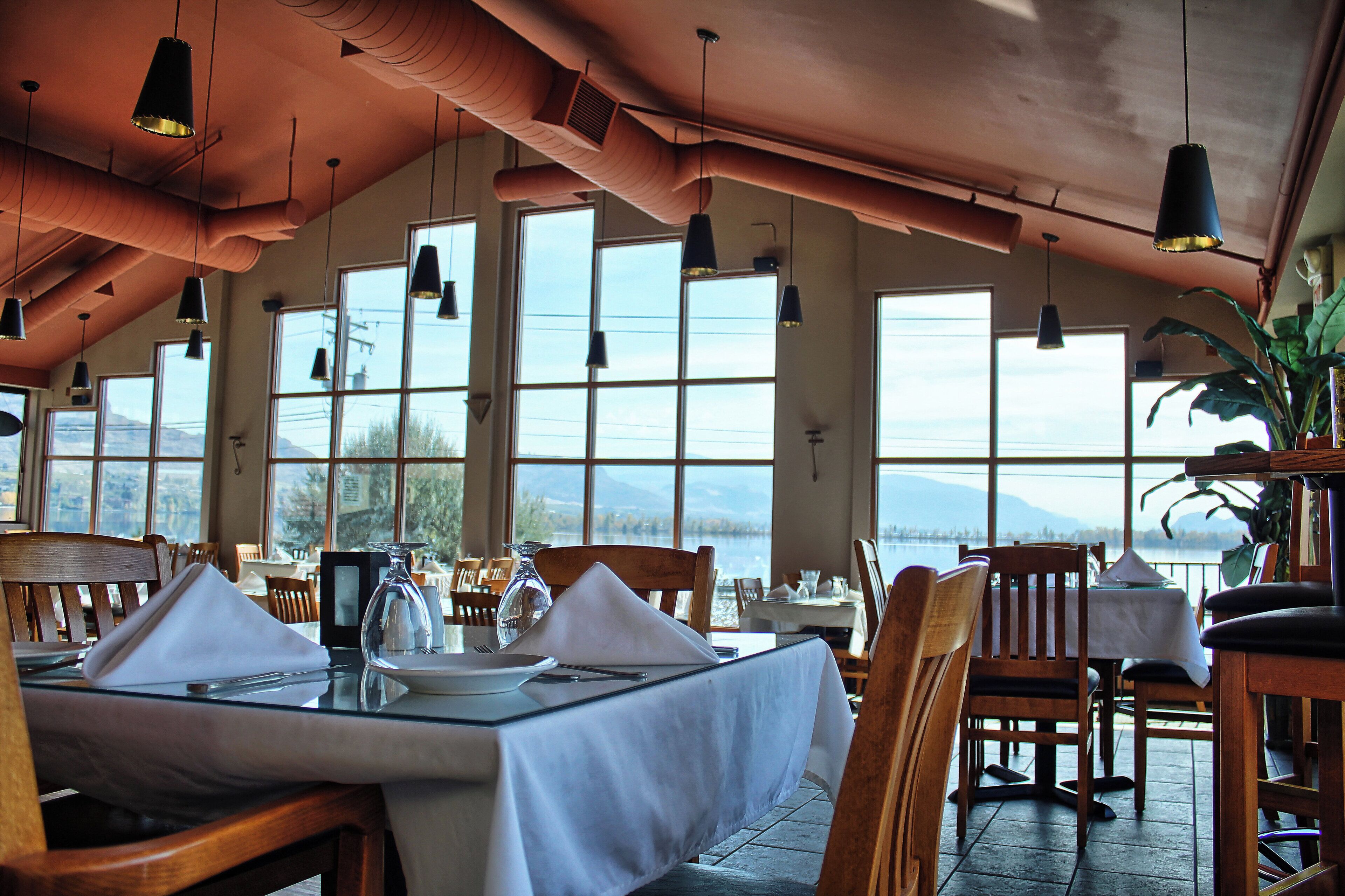 A restaurant with a high ceiling, large windows, and a view of a lake. Tables are set with white tablecloths and silverware.