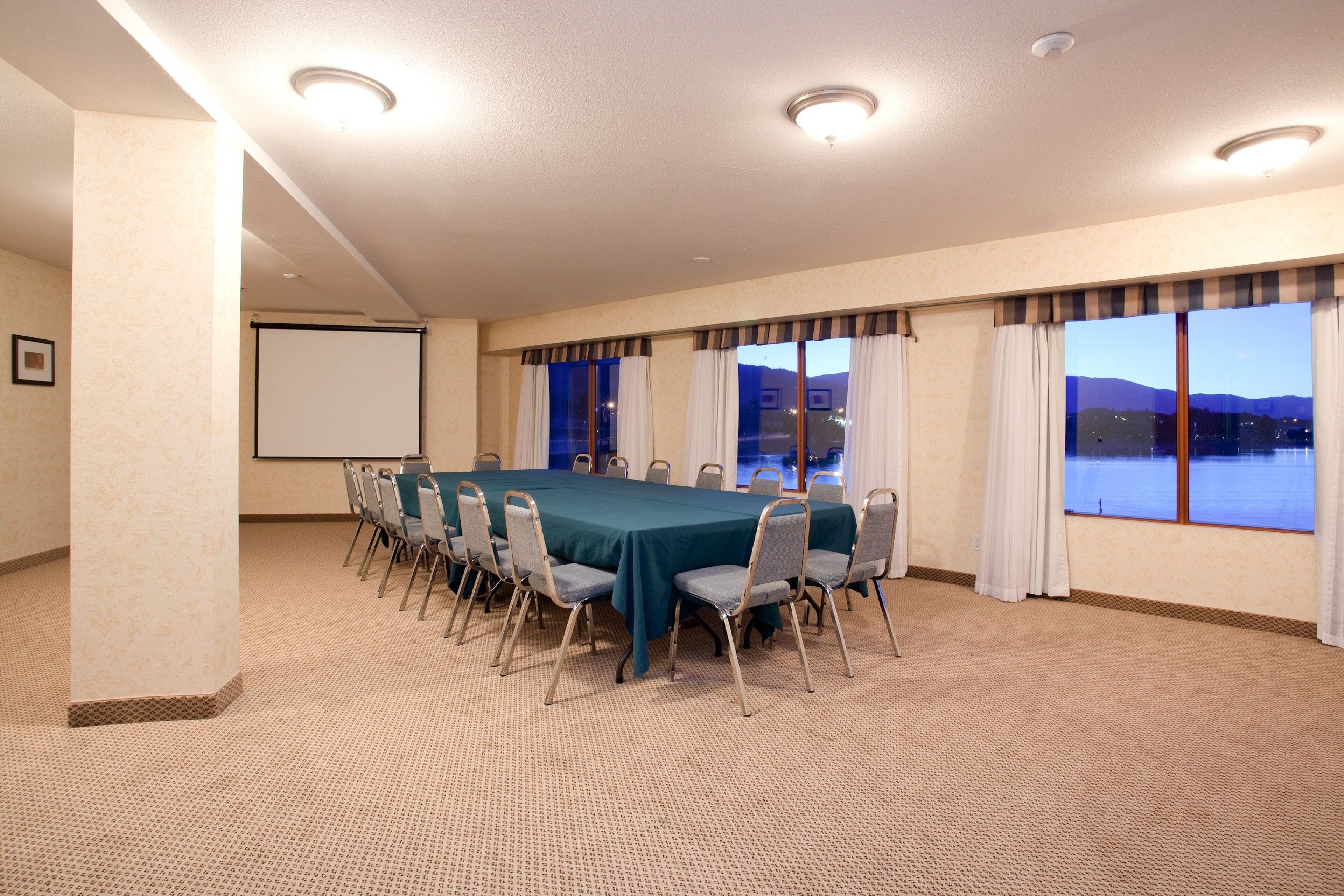 A conference room with a large table, chairs, a projector screen, and windows with curtains.