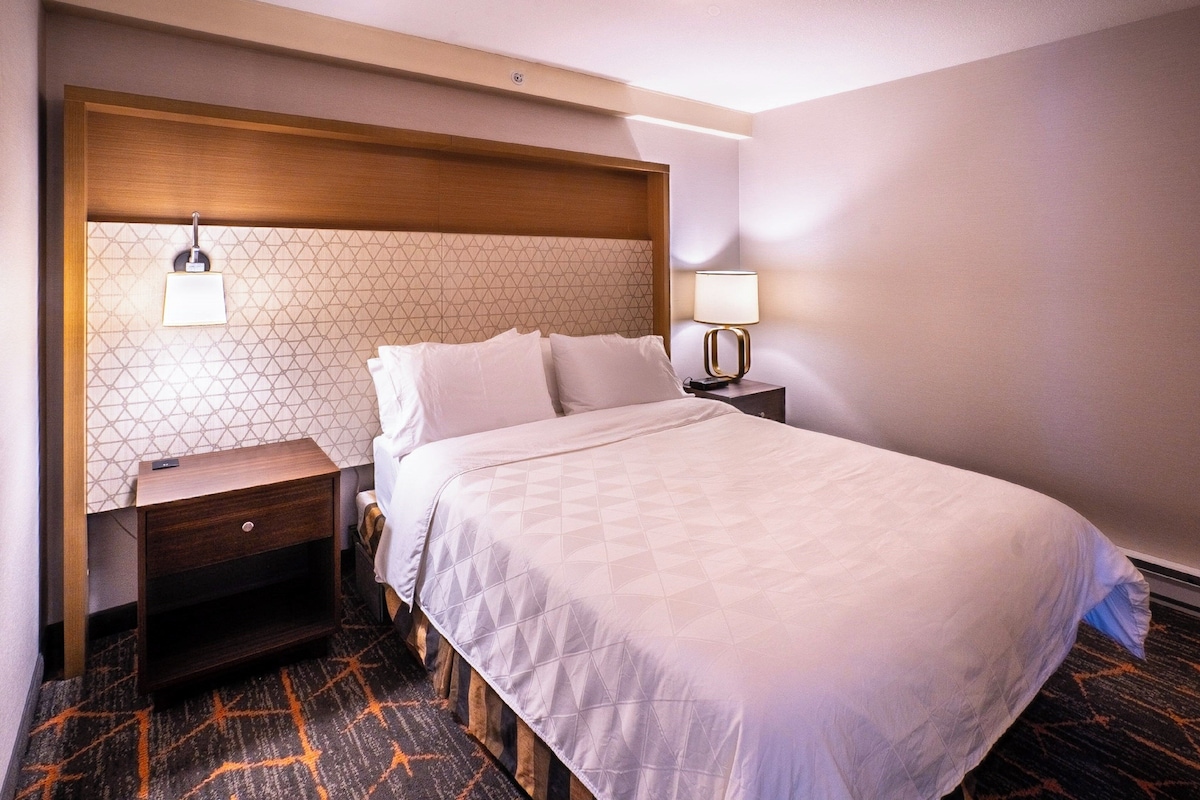 Une chambre d’hôtel avec un grand lit, une tête de lit en bois, une table de chevet, une lampe et une moquette à motifs.