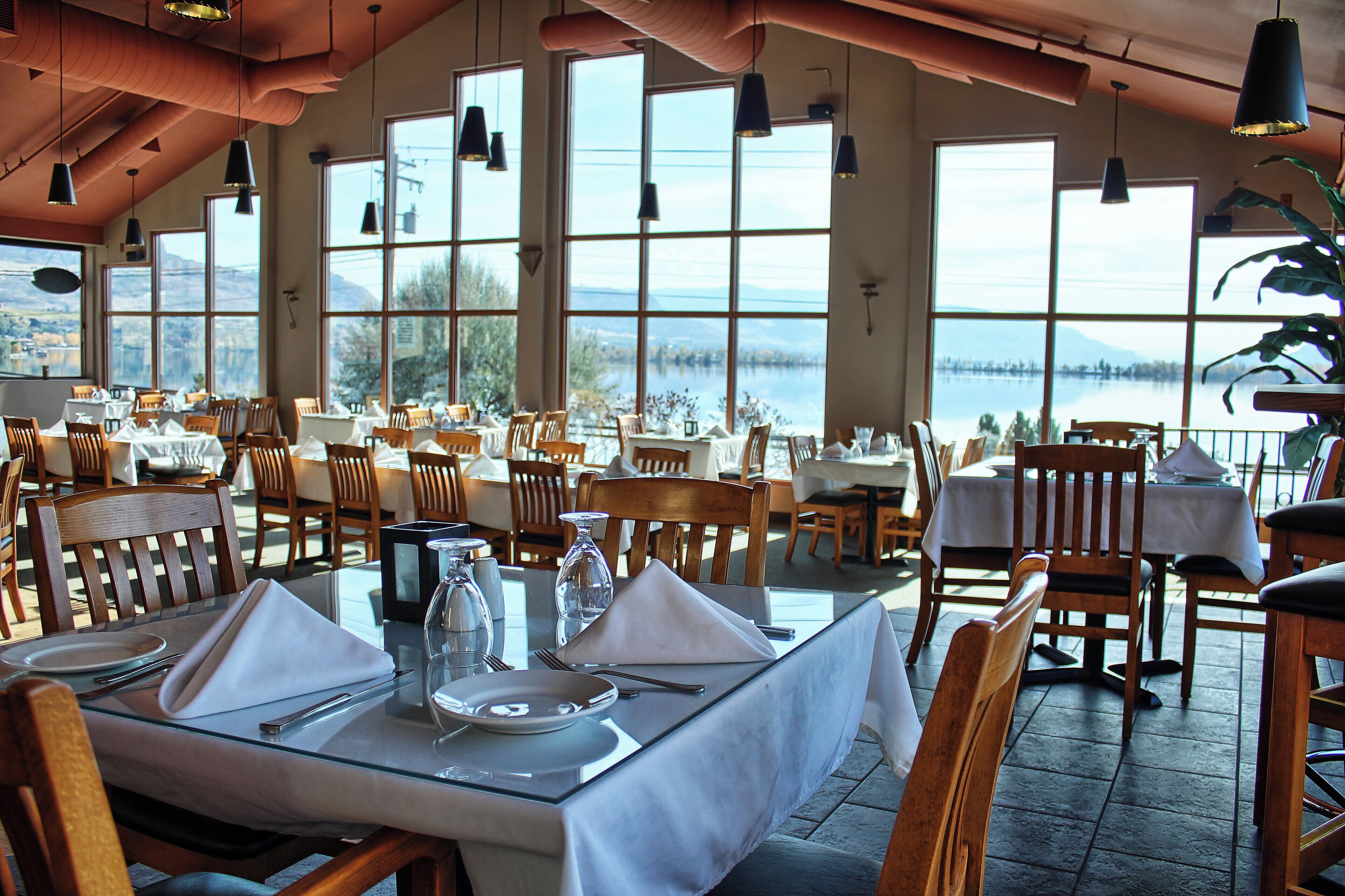 Un restaurante con mesas dispuestas para comer, amplias ventanas que dan a un lago y lámparas colgantes.