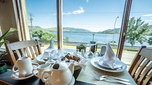 A dining table set with white tableware, a teapot, and a basket of snacks, overlooking a lake with mountains in the background.