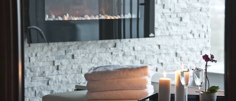 Zona de spa con pared de ladrillo blanco, chimenea, una pila de toallas, velas y una pequeña planta en maceta.