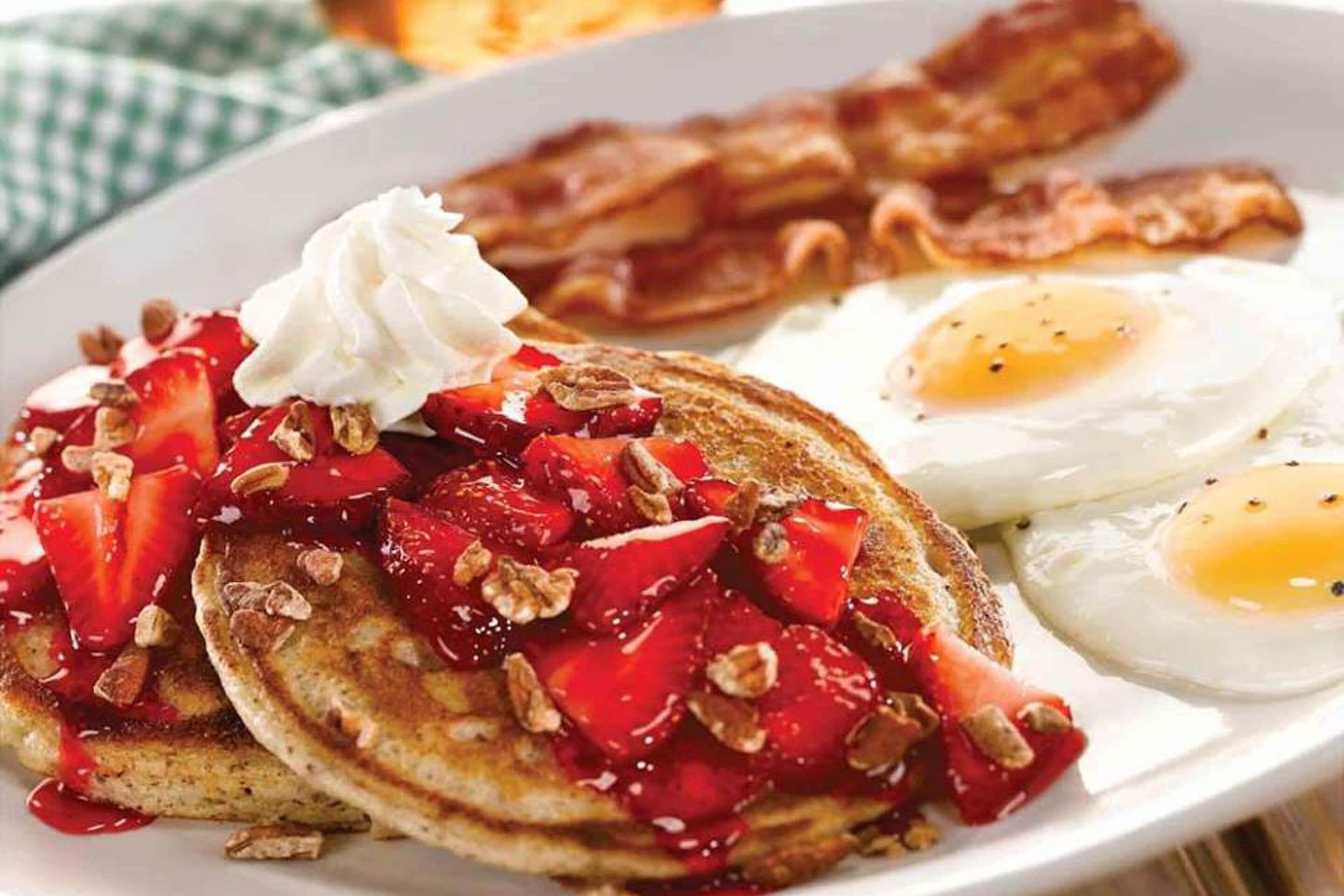 A plate of pancakes topped with strawberries, whipped cream, and granola, accompanied by two fried eggs and bacon strips.
