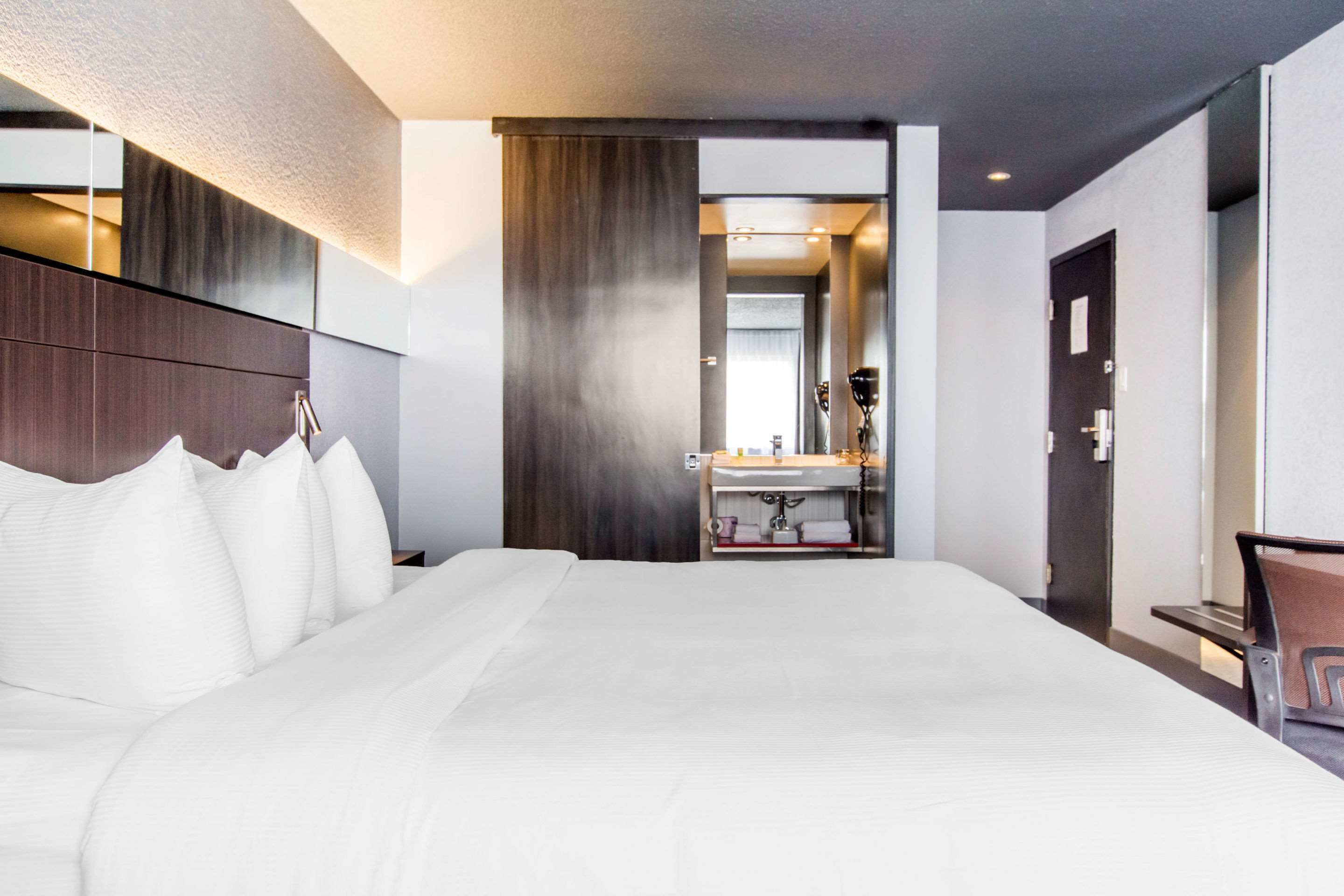 A hotel room with a large bed, a wooden headboard, a bathroom with a sink and mirror, and a door leading to another room.