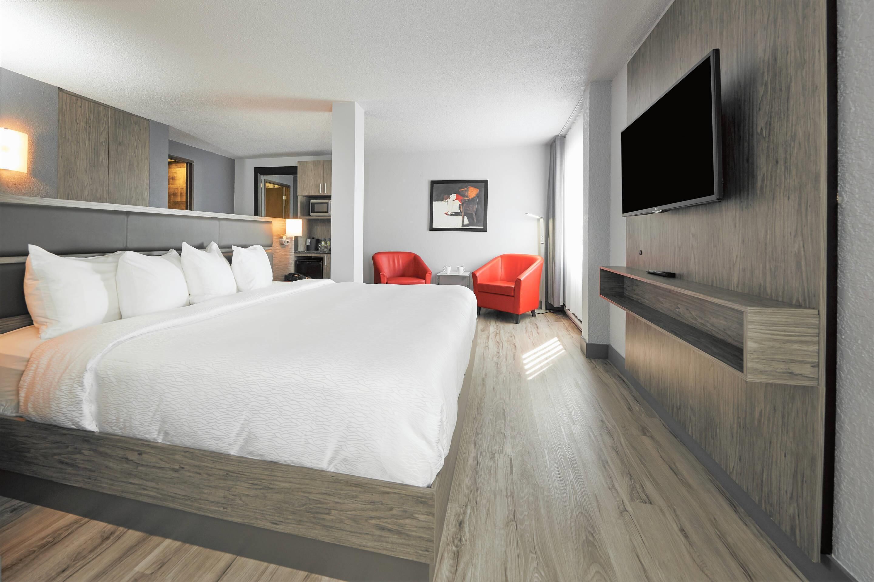 A modern hotel room with a large bed, a flat-screen TV mounted on the wall, and a small seating area with two red chairs.