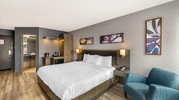 A hotel room with a large bed, a blue armchair, a desk with a lamp, and artwork on the walls.