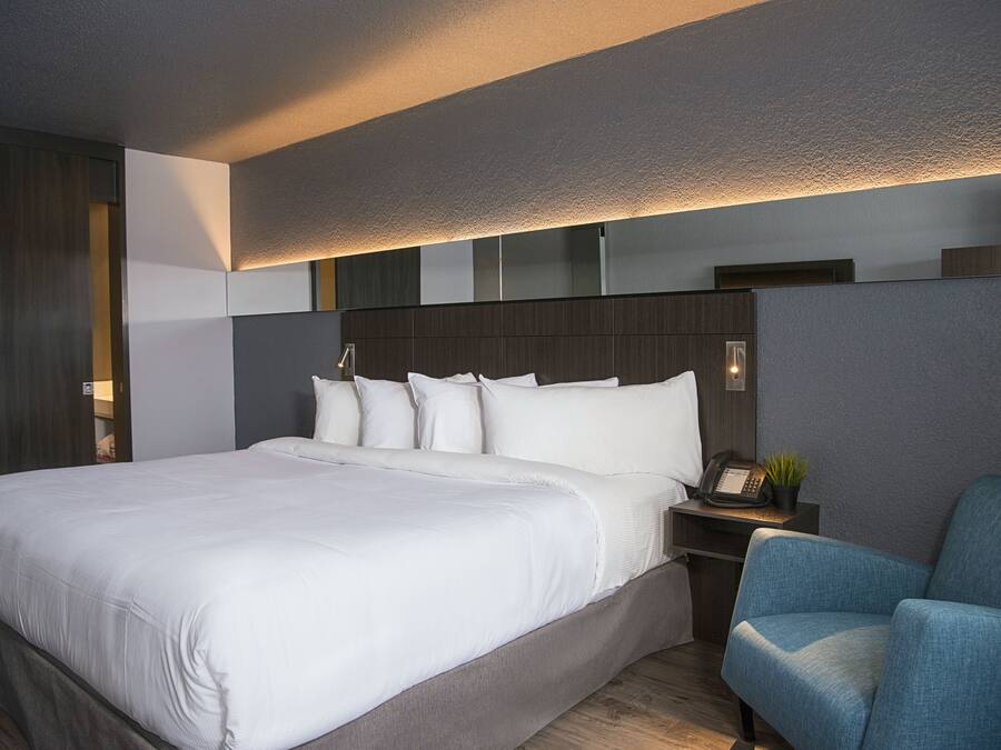 A modern hotel room with a large bed, a desk with a telephone, and a blue armchair.