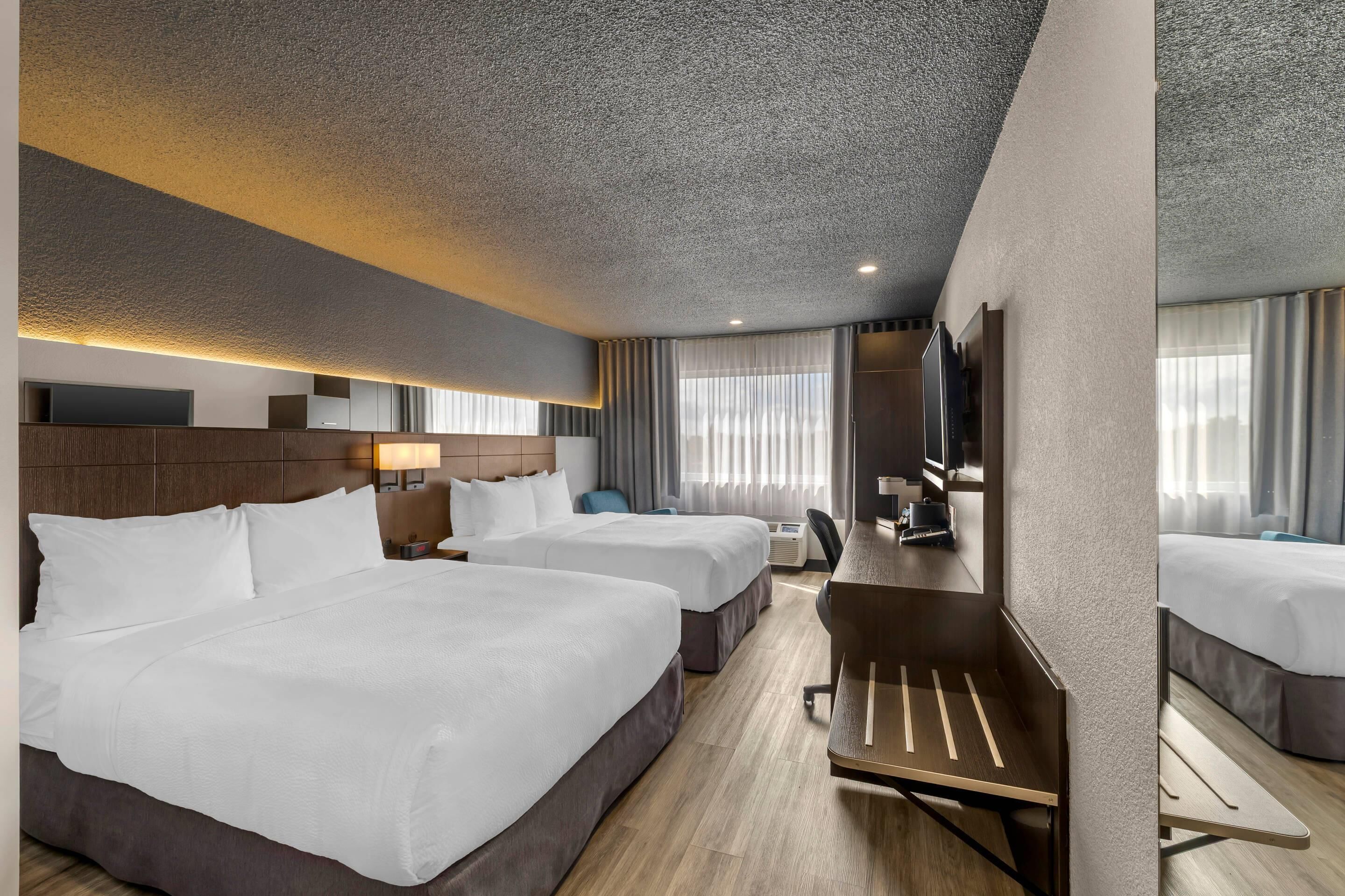 Habitación de hotel con dos camas, un escritorio, una silla, un televisor y un espejo grande.