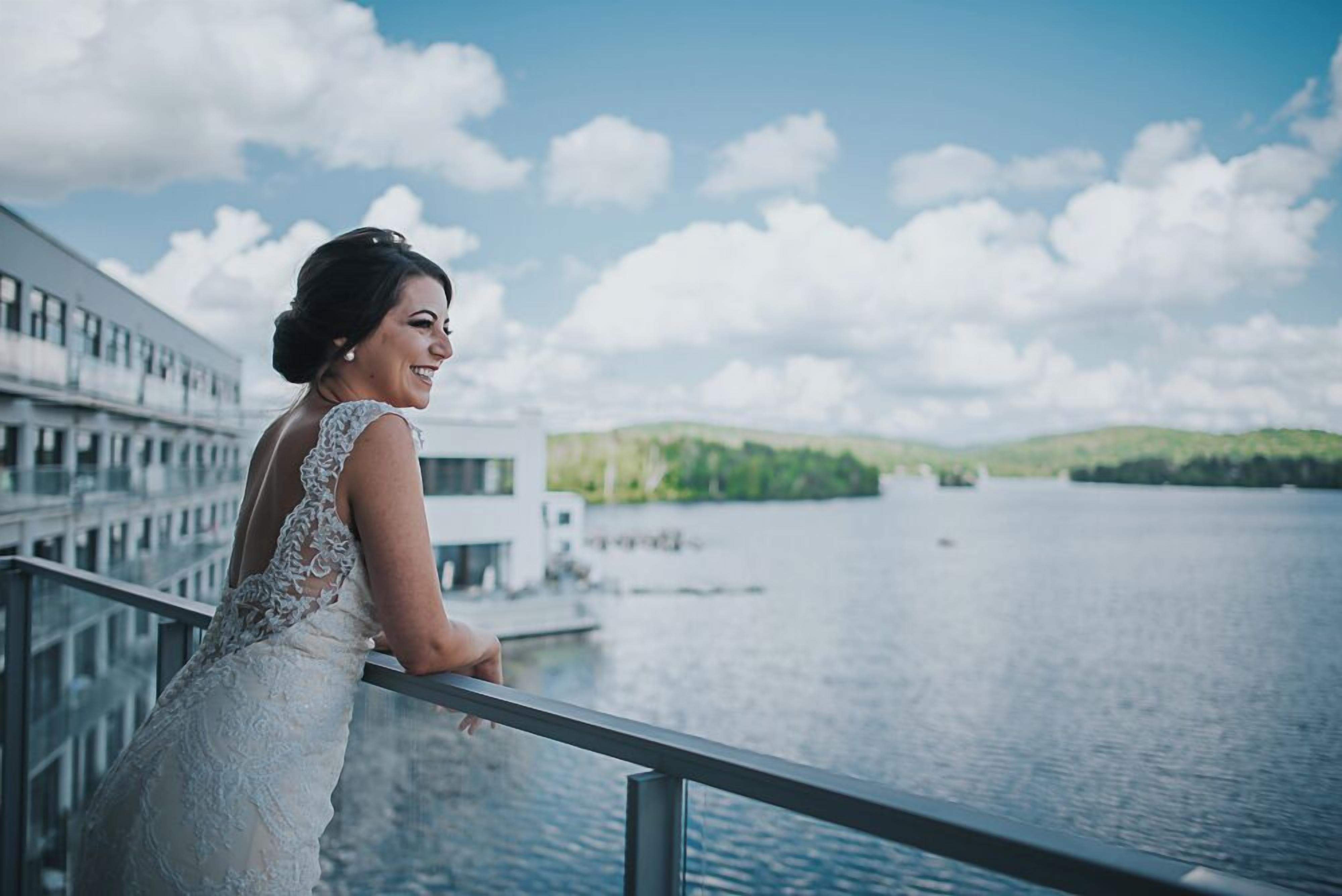 Una persona con vestido blanco apoyada en la barandilla de un balcón, con vistas a una masa de agua y edificios al fondo.
