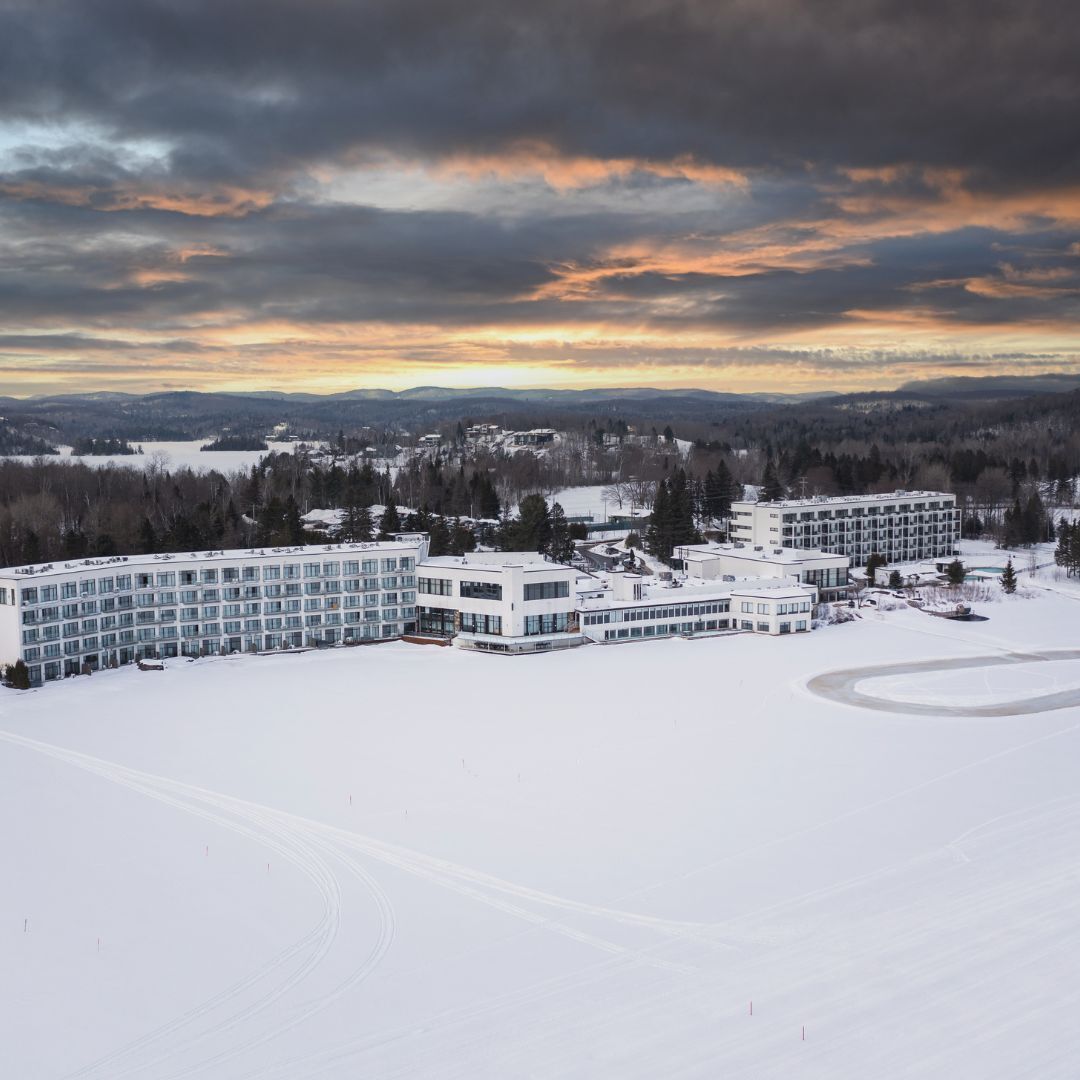 A snow-covered hotel complex with multiple buildings, a large parking area, and a scenic view of mountains and a lake under a cloudy sky.