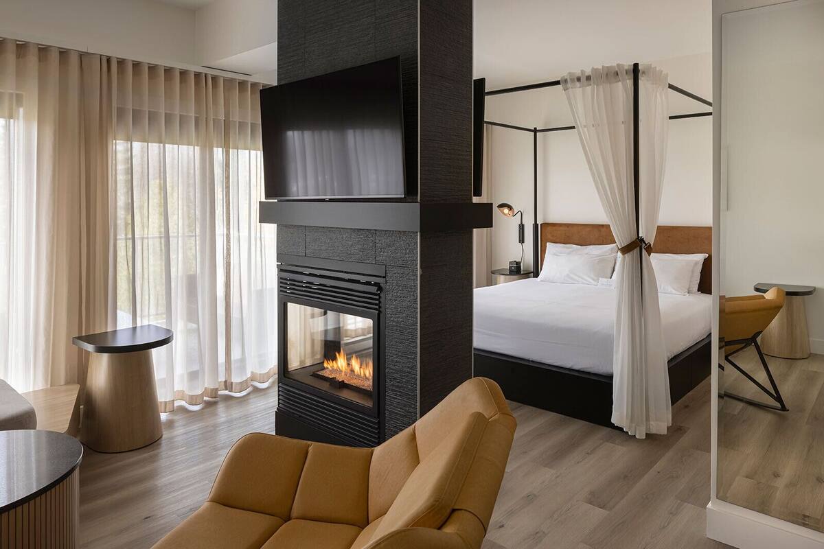 A modern hotel room with a large bed, a fireplace, a sofa, and a dining area.